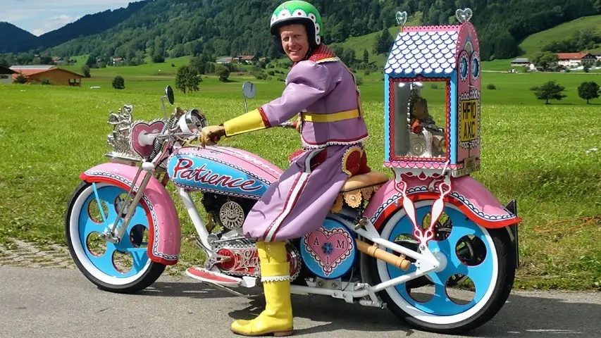 Grayson Perry’s on his pink motorbike carrying his teddybear and childhood hero, Alan Measles, in a glass shrine on the back.